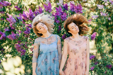 Two beautiful lifestyle excited sisters have fun outdoor in lilac bushes. Twins young happy smiling girls models hold hands and shaking heads. Similar appearance women childhood behavior at nature.