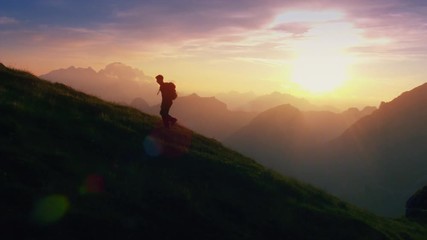 aerial - epic shot of a man hiking on the edge of the mountain as a silhouette in colorful sunset (e