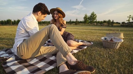 Poster - Young happy loving couple sitting outdoors on the picnic in the field drinking wine talking with each other