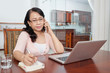 © DragonImages - Attractive mature Asian woman sitting at home table with laptop, talking on smartphone and writing notes in notepad