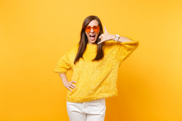 Woman in heart orange glasses doing phone gesture like says: call me back with hand and fingers like talking on the telephone isolated on yellow background. Communicating, people emotions, lifestyle.