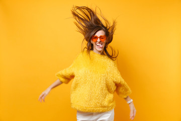 Comic young girl in fur sweater heart orange glasses showing tongue, fooling around in studio jump with flying hair isolated on yellow background. People sincere emotions, lifestyle. Advertising area.