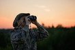 The caucasian man is wearing a camouflage suit watching through binoculars in outdoors. The birdwatcher with field glasses is in a twilight in rural.