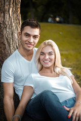 Happy young cute loving couple sitting in park near tree looking at camera hugging.