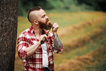 Wall Mural - man brutal lumberjack with beard and moustache in red checkered shirt shaves with axe blade on natural background