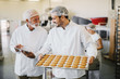 © dusanpetkovic1 - Picture of two employees in sterile clothes in food factory smiling and talking. Younger man is holding tray full of fresh cookies while the older is holding tablet and checking production line.