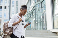 Happy Relaxed Young African Female Student In White Basic Shirt And Trendy Sunglasses With Leather Backpack Looking At Her Smartphone In Summer Urban Street Over Modern Business Centre Buildings.