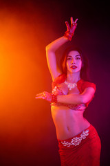 Wall Mural - Tempting sexy traditional oriental belly dancer girl dancing on purple neon smoke background. Woman in exotic red costume sexually moves her semi-nude body.