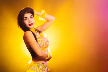 Wall Mural - Alluring sexy traditional oriental belly dancer girl dancing on yellow neon smoke background. Woman in exotic costume with feathers sexually moves her semi-nude body