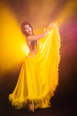 Wall Mural - Seductive sexy traditional oriental belly dancer girl dancing on yellow neon smoke background. Woman in exotic costume with feathers sexually moves her semi-nude body.