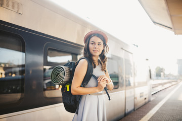 theme railway and travel. Portrait young caucasian woman with toothy smile standing at train station train background with backpack and equipment for tourism isolated mat in dress and hat in summer