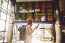 Theme Travel And Tranosport. Beautiful Young Caucasian Woman In Dress And Backpack Standing Inside Train Station Or Terminal Looking At A Schedule Holding A Red Phone, Uses Communication Technology