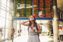 Theme Travel And Tranosport. Beautiful Young Caucasian Woman In Dress And Backpack Standing Inside Train Station Or Terminal Looking At A Schedule Holding A Red Phone, Uses Communication Technology