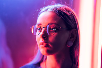 Wall Mural - Millennial enigmatic pretty girl with unusual dyed hairstyle near glowing neon wall at night. Blue hair, golden sequins as freckles,nose piercing. Mysterious hipster teenager in glasses