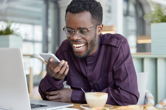 Dark skinned graphic designer sends text message, glad to recieve good notification, sits at desktop, drinks hot coffee, dressed in purple shirt. Handsome black man plans his freelance schedule
