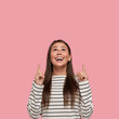 © WHstudio Leushin N - Isolated shot of happy girlfriend being in high spirit, points with both index fingers upwards, dressed in striped casual sweater, poses over pink background. Hey, look upside, please. Advertisement