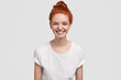 Adorable freckled young lady or teenager smiles joyfully at camera, has red hair combed in knot, dressed at casual t shirt in one tone with background, thinks about good things. Emotions concept