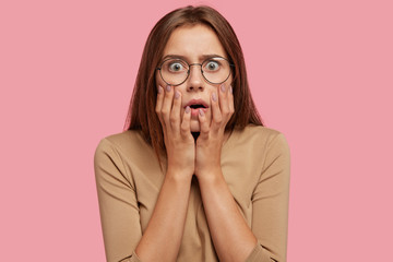 Wall Mural - Half length portrait of beautiful female being very emotional and surprised, stares with bugged eyes, has stupor, dressed casually, isolated over pink background. Amazement and fear concept.