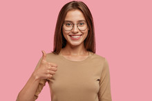 Portrait Of Happy Teenager Keeps Thumb Raised, Being In Good Mood, Shows Her Agreement, Poses Over Pink Background, Says: Nice Joke! Young Woman Shows Like Gesture, Satisfied With Something.