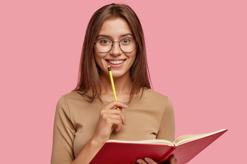 Photo of happy brunette woman with positive smile, carries textbook, holds pencil for writing, makes notes while listens some information, poses over pink background. Its time for creating new poem!
