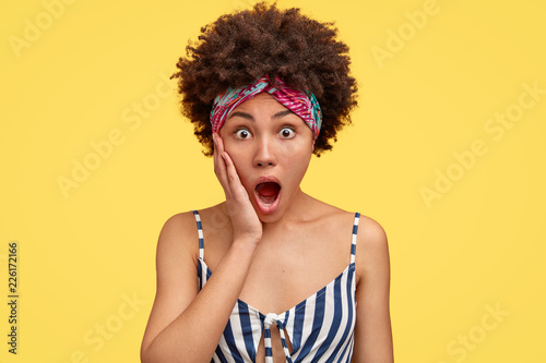 Fotografía Shocked housewife witnessed terrible crime or accident, holds palm near widely opened mouth, has Afro hairstyle, dressed in casual striped t shirt, isolated over yellow background