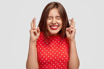 Wall Mural - Pleased attractive European woman makes wish to win, raises hands with crossed fingers, waits for lottery results, closes eyes, has red lips, dressed in fashionable dress, isolated over white wall
