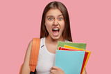 Fototapeta  - Outraged brunette woman exclaims with fury, keeps mouth widely opened, feels irritated with lazy groupmate, models against pink studio wall, holds textbooks or notepads, has discontent expression