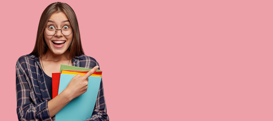 Horizontal view of happy dark haired woman with toothy smile, express positive emotions, cant catch breath from amazement, wears round glasses, points with index finger at copy space for your slogan