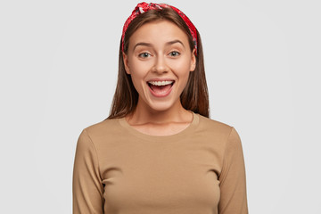 Happy green eyed European woman with pleased expression, keeps mouth opened, wears red bandana, beige casual sweater, being in good mood as spends leisure time in family circle, stands indoor.