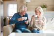 © fizkes - Happy senior old couple sitting on sofa in living room talking enjoying tea at home, smiling middle aged mature family chatting laughing drinking coffee having fun pleasant conversation together