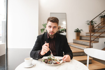 Handsome business man with a beard sitting in a cozy restaurant at the table, eating salad and looking at the camera. Portrait of a business man at breakfast in the bright café.