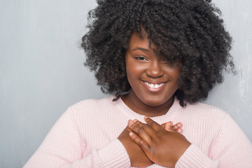 Young african american plus size woman over grey grunge wall wearing winter sweater smiling with hands on chest with closed eyes and grateful gesture on face. Health concept.