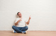 © Krakenimages.com - Young caucasian man sitting on the floor over white brick wall smiling and looking at the camera pointing with two hands and fingers to the side.