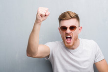 Young Redhead Man Over Grey Grunge Wall Wearing Retro Sunglasses Annoyed And Frustrated Shouting With Anger, Crazy And Yelling With Raised Hand, Anger Concept