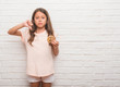 © Krakenimages.com - Young hispanic kid over white brick wall eating chocolate chips cooky with angry face, negative sign showing dislike with thumbs down, rejection concept