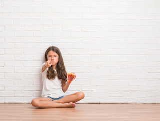 Poster - Young hispanic kid sitting on the floor over white brick wall eating pizza slice pointing with finger to the camera and to you, hand sign, positive and confident gesture from the front