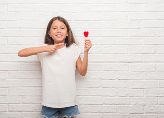 Poster - Young hispanic kid over white brick wall eating red heart lollipop candy very happy pointing with hand and finger