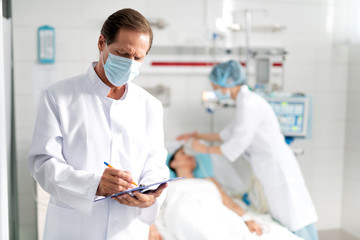 Waist up portrait of male doctor in protective mask writing down information about condition of young lady on clipboard. Female assistant doing artificial inhalation on blurred background