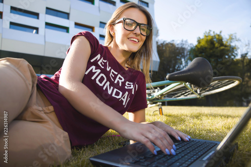 Fotografie, Obraz Pretty student woman wearing glasses lying near the university and typing on the