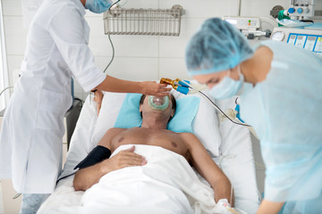 Unconscious middle aged man getting mechanical ventilation in hospital. Cropped female medical worker using respiratory equipment