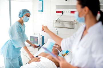 Female medical worker in protective mask and cap taking care of sick middle aged man. Blurred young doctor pointing at monitor with indications of heart rate