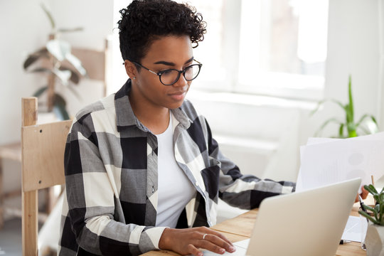 Millennial young african employee in eyeglasses hold documents papers with charts and graphics using computer sitting at the desk in office room or home feels confident, fruitful working day concept