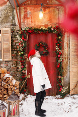  Young smiling teenage girl in white coat and red hat. Outdoor winter portrait of  blonde teen girl in red glasses. Chrismas wreath and red painted wooden door at background.