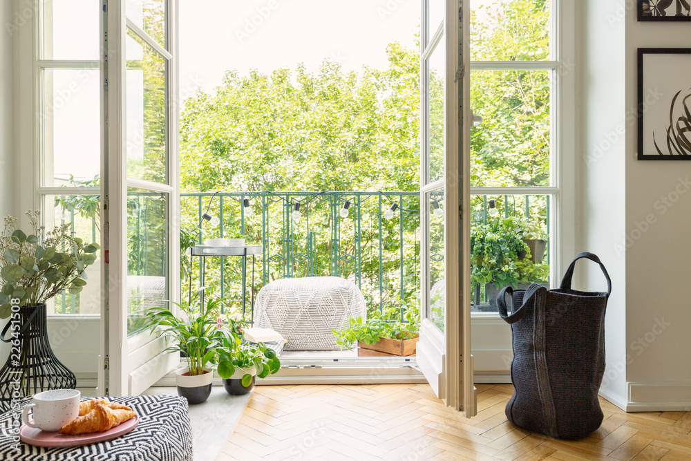Open glass door from a living room interior into a city garden on a sunny balcony with green plants and comfy furniture - obrazy, fototapety, plakaty 