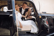Look from the outside at adorable wedding couple in classy dresses sitting inside a black retro car and hugging each other tender
