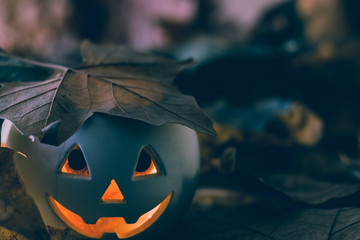 White Halloween pumpkin with dark eyes and a hat leaf illuminated with orange light among autumnal leaves