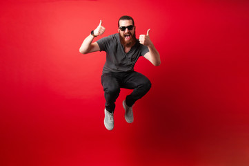 Cheerful bearded hipster man with sunglasses jump over red background and showing thumbs up