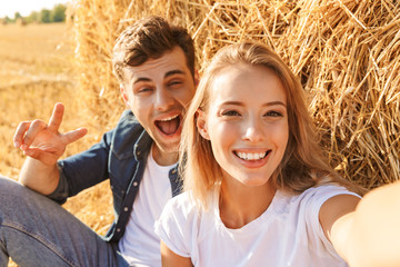 Wall Mural - Photo of cheerful couple man and woman taking selfie while sitting under big haystack in golden field, during sunny day