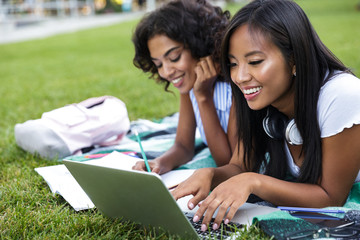 Wall Mural - Two cheerful young girls students sitting on a grass