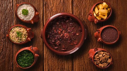 Wall Mural - traditional Brazilian feijoada in stop motion with portions of cassava, bacon, kale, flour, rice and sauce. Top view with old wood background.